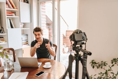 cheerful vlogger pointing with fingers at digital camera while sitting near laptop clipart