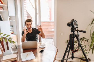 neşeli vlogger dizüstü bilgisayarın yanında otururken ve tripoddaki dijital kameraya bakarken iyi jestler gösteriyor