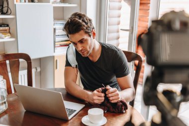 Kulağında tıbbi maskeyle dijital kameranın yanında örgü örerken laptopa bakan yoğunlaştırılmış vlogger odaklı