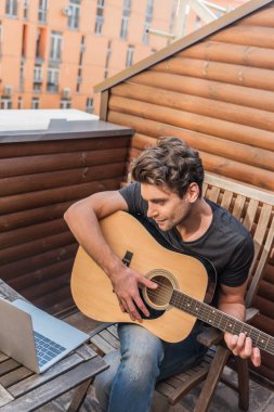 Balkonda oturup dizüstü bilgisayara bakarken gitar çalan genç adamın yüksek açılı görüntüsü