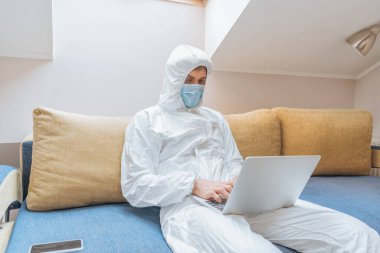 Tehlikeli madde giysisi ve koruyucu maskeli genç adam akıllı telefonun yanındaki koltukta oturuyor ve bilgisayarını kullanıyor.