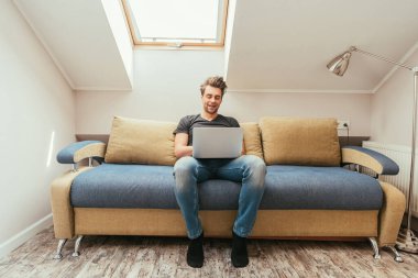 Gülümseyen genç adam evdeki kanepede otururken dizüstü bilgisayar kullanıyor.
