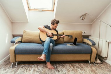 Genç adam kanepede oturup boş ekranlı dizüstü bilgisayara bakarken gitar çalıyor.