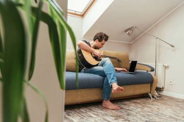 Genç bir adamın gitar çalarken dizüstü bilgisayarın yanında boş ekranla oturduğu seçici odak noktası.