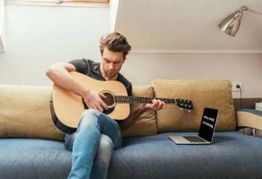 Bilgisayarın yanındaki koltukta otururken gitar çalan konsantre genç adam ekranda online öğrenme yazısıyla