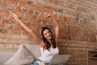 Heyecanlı bir kadın, kulaklık ve dizüstü bilgisayarla kendini tecrit sırasında müzik dinliyor.