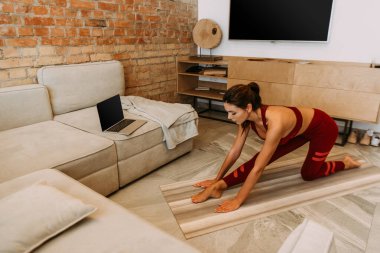 Çekici bir kız internette dizüstü bilgisayarla yoga minderinin üzerinde kendini izole ediyor.