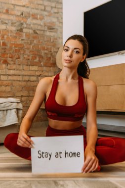 beautiful sportswoman holding stay at home sign while sitting on fitness mat at home during self isolation clipart