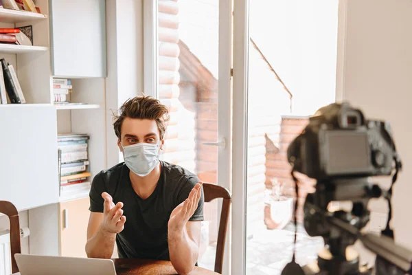 デジタルカメラを見ながら医療用マスクジェスチャーの若いビデオブロガーの選択的な焦点 — ストック写真