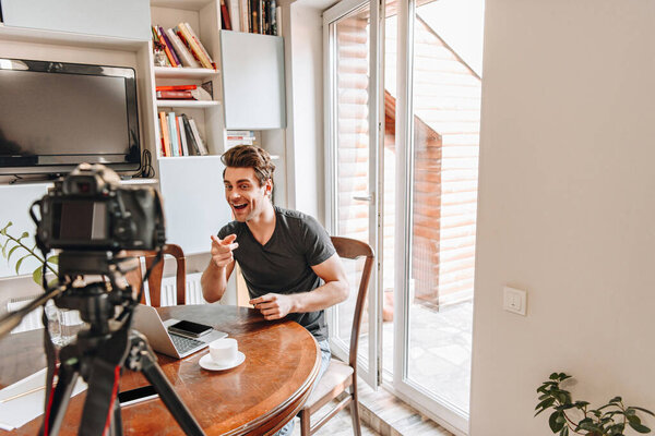 selective focus of cheerful vlogger pointing with finger at digital camera on tripod