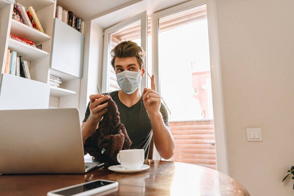 молодой человек в медицинской маске показывает жест идеи во время вязания рядом с ноутбуком
