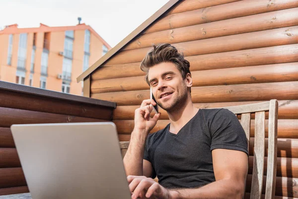 英俊男子坐在阳台上 一边用笔记本电脑 一边用智能手机交谈 一边对着相机微笑 — 图库照片