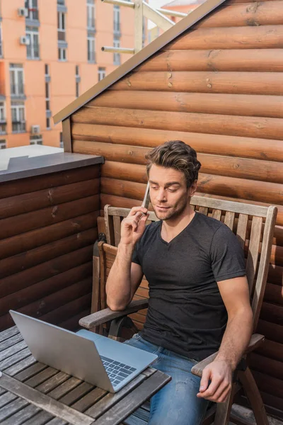 Junger Skeptischer Mann Nutzt Laptop Und Telefoniert Auf Dem Balkon — Stockfoto