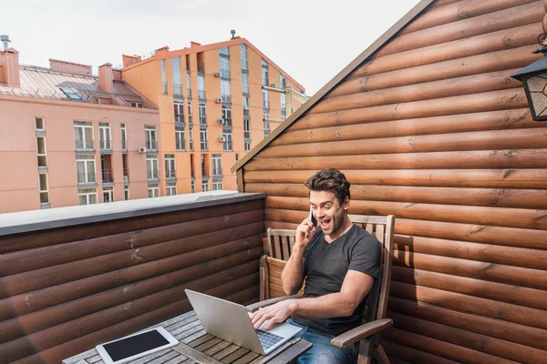 Excited Man Using Laptop Talking Smartphone While Sitting Balcony — Stock Photo, Image