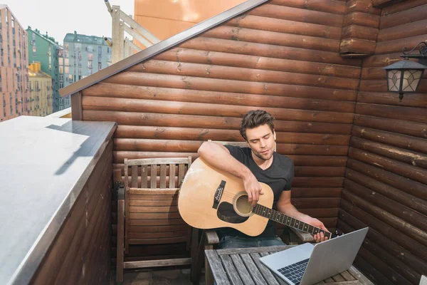 Gitar tutan ve balkonda otururken dizüstü bilgisayara bakan genç adam.