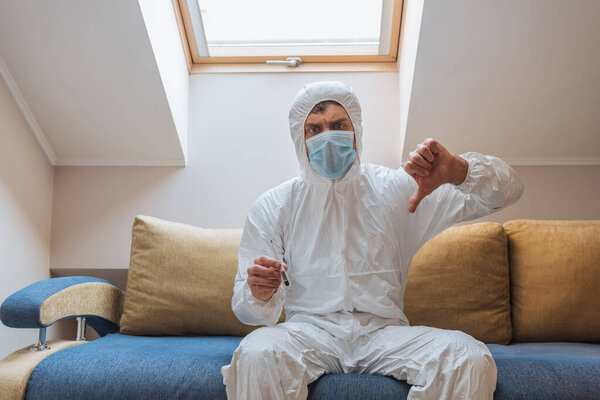 displeased man in hazmat suit and protective mask showing thumb down while holding thermometer