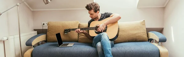 年轻男子坐在沙发上弹奏吉他 看着屏幕空白的笔记本电脑的水平图像 — 图库照片