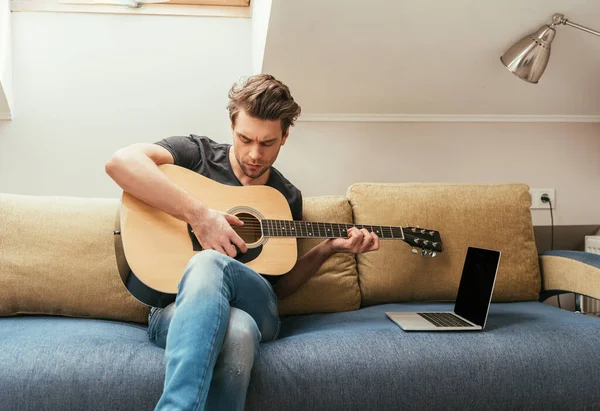 集中注意力的年轻人坐在沙发上弹奏吉他手 沙发靠近笔记本电脑 屏幕空白 — 图库照片