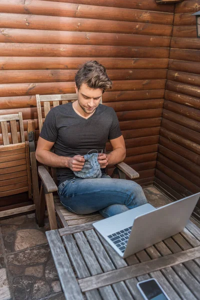 High angle view of handsome young man knitting while sitting near laptop on balcony — Stock Image High Angle View Handsome Young Man Knitting While Sitting Laptop Stock Image