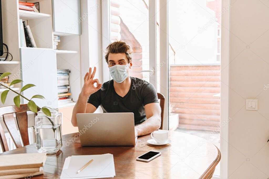young man in protective mask showing thumb down near laptop and cup of coffee on table