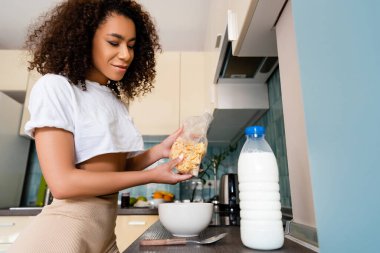 Afro-Amerikalı bir kadın elinde mısır gevreğiyle kasenin yanında süt şişesi tutuyor. 