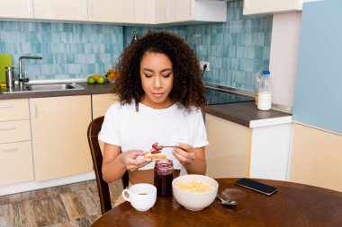 attractive african american woman holding spoon with sweet jam near toast bread and smartphone with blank screen  clipart