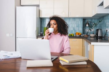 Kıvırcık Afrikalı Amerikalı serbest yazar laptopun, kitapların ve belgelerin yanında kahve içiyor. 