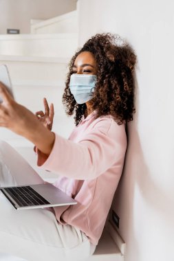 Seçici odak noktası, tıbbi maskeli Afro-Amerikan serbest yazar merdivenlerde oturuyor ve dizüstü bilgisayarın yanında selfie çekerken barış işareti gösteriyor. 