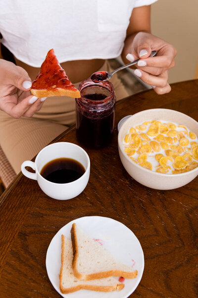 cropped view of african american woman holding spoon with jam near toast bread, corn flakes in bowl and cup with coffee on table 