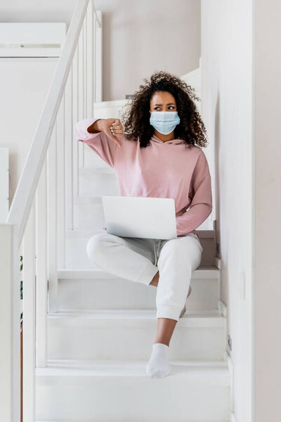 curly african american freelancer in medical mask sitting on stairs and showing thumb down near laptop 