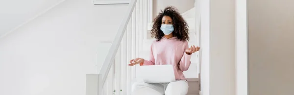 panoramic concept of confused african american freelancer in medical mask sitting on stairs and showing shrug gesture near laptop