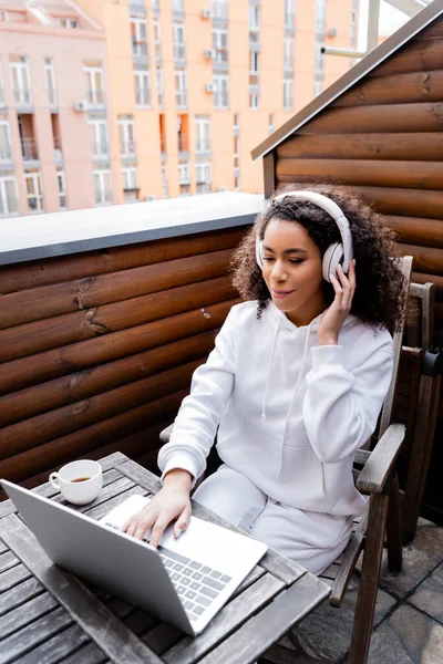 Rizado Afroamericano Freelancer Tocando Auriculares Inalámbricos Utilizando Ordenador Portátil Cerca —  Fotos de Stock