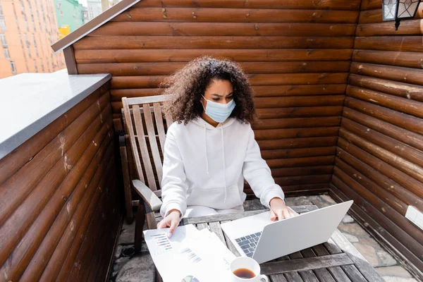 アフリカ系アメリカ人の医療用マスクのフリーランスは コーヒー チャート グラフのカップの近くにノートパソコンを使って — ストック写真