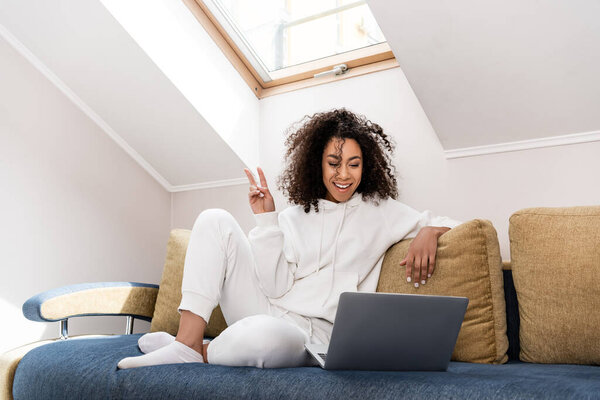 happy african american girl sitting on sofa and showing peace sign near laptop while having video call 