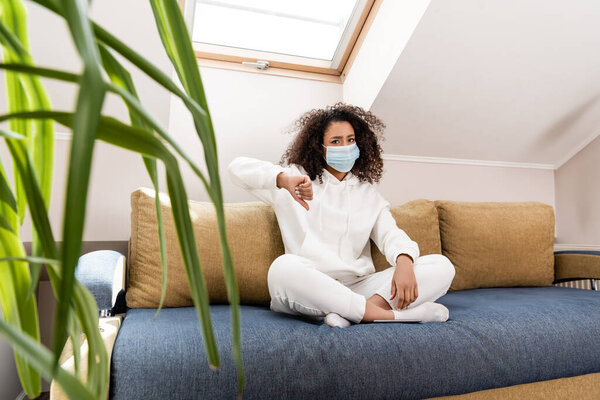 selective focus of curly african american girl in medical mask showing thumb down while sitting on sofa 