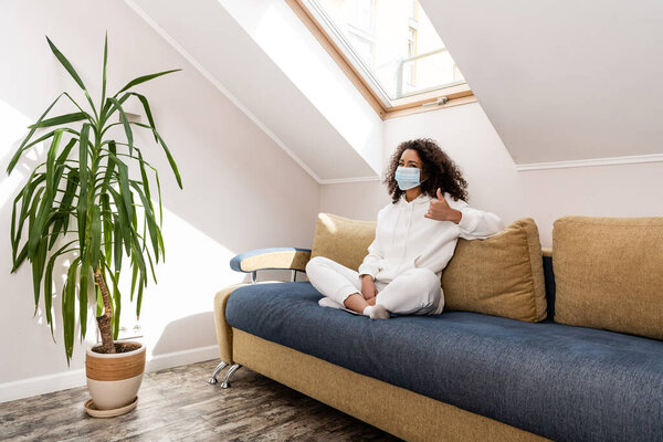 curly african american girl in medical mask showing thumb up while sitting on sofa 
