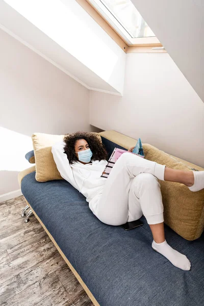 Afrykańska Amerykanka Masce Medycznej Leżąca Kanapie Czytająca Magazyn — Zdjęcie stockowe