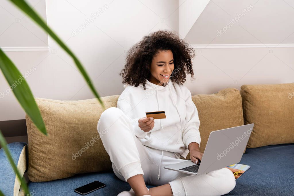 selective focus of happy african american girl holding credit card near laptop while sitting on sofa