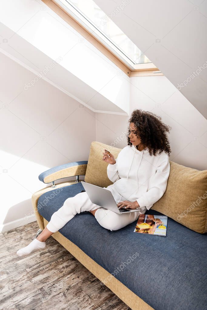 curly african american girl holding credit card near laptop while sitting near magazine on sofa