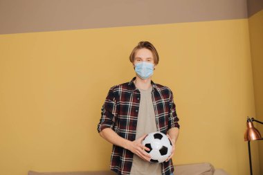 Medikal maskeli genç adam futbol topu tutuyor ve kameraya bakıyor.
