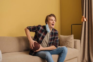 Sağlık maskeli heyecanlı adam oturma odasında şampiyonluk maçı izlerken elinde Amerikan futbolu tutuyor.