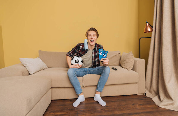 excited man in medical mask holding football and sport match tickets near remote controller, end of quarantine concept