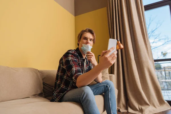 Geschokte Man Medisch Masker Zoek Naar Smartphone Woonkamer — Stockfoto