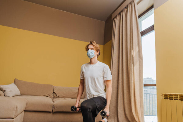 man in medical mask exercising with dumbbells in living room