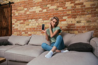 Kanepede otururken akıllı telefonu boş ekranla gösteren güzel bir genç kadın.