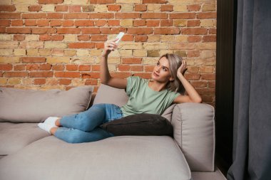 attractive young woman taking selfie on smartphone while resting on sofa near brick wall clipart