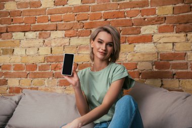 Kanepede oturmuş kameraya gülümseyen ve boş ekranlı akıllı telefonu gösteren güzel bir kadın.