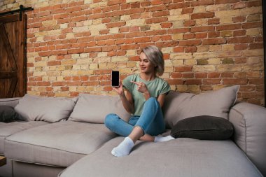 Çekici genç bir kadın kanepede oturuyor ve boş ekranlı akıllı telefonu işaret ediyor.
