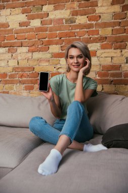 Tuğla duvarın yanındaki kanepede oturan mutlu genç kadın ve boş ekranlı akıllı telefonu gösteriyor.