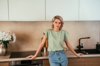 Ciddi, çekici bir kadın mutfakta kameraya bakıyor.